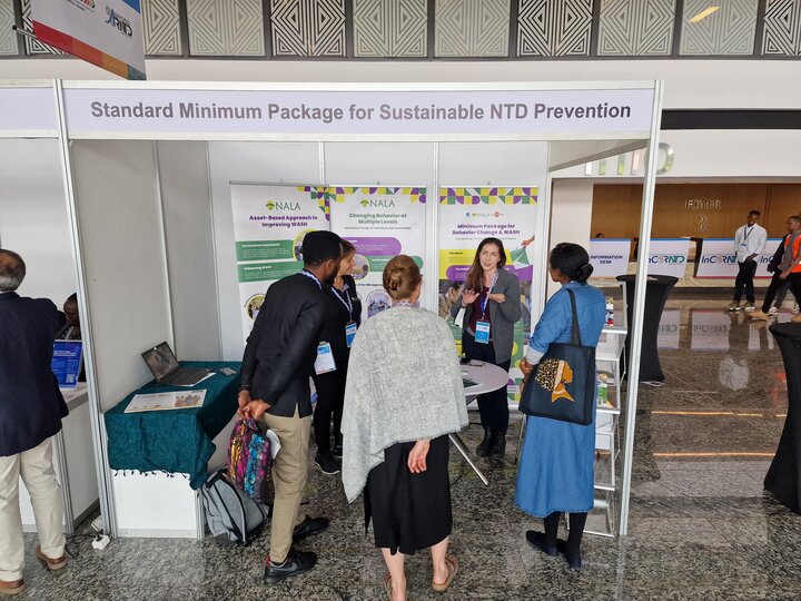 Naomi Caplan standing in front of a display talking to a group of people about NALA's Standard Minimum Package for Sustainable NTD Prevention. 